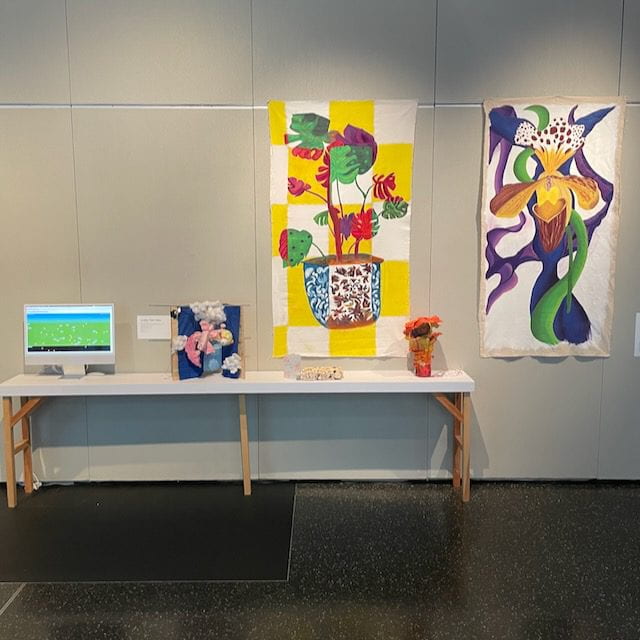 Displayed on a long white table are a computer monitor featuring a waste-collecting game, an animatronic device, a mini-zine and some blocks of organic and fungi texture, and an orange mushroom sculpture. Above and to the right of the table are two large-scale canvas depicting flowers.