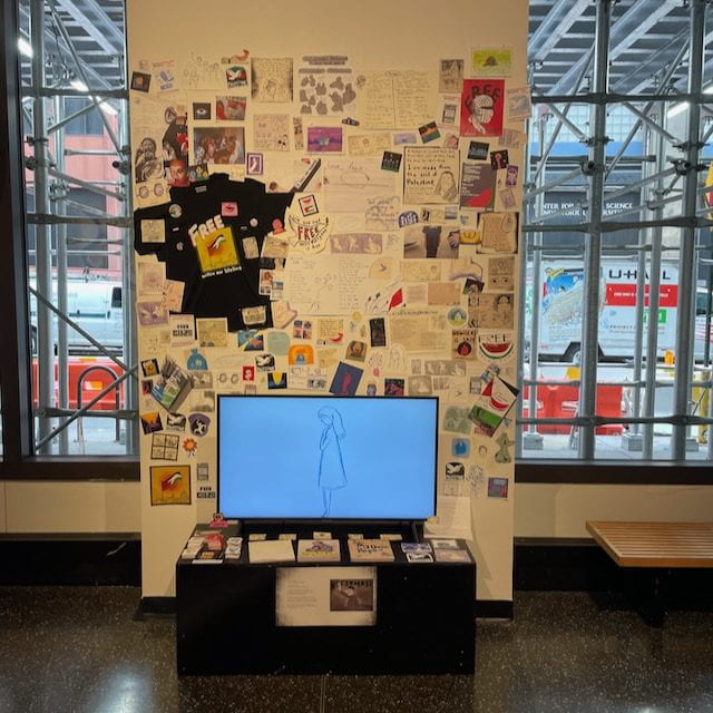 A section of the wall, flanked by large windows that shows views of the street and scaffolding, is covered by posters, stickers, photos, zines of various sizes. The words "FREE PALESTINE" and similar slogans are visible on many displayed objects, including a black t-shirt with a print of Palestine's national flag. In front of the wall is a TV set on a black pedestal, placed horizontally. On the TV screen is the drawn outline of a woman in profile, putting her hands on her chest. Stickers, artist's books, and more prints are placed on the pedestal.