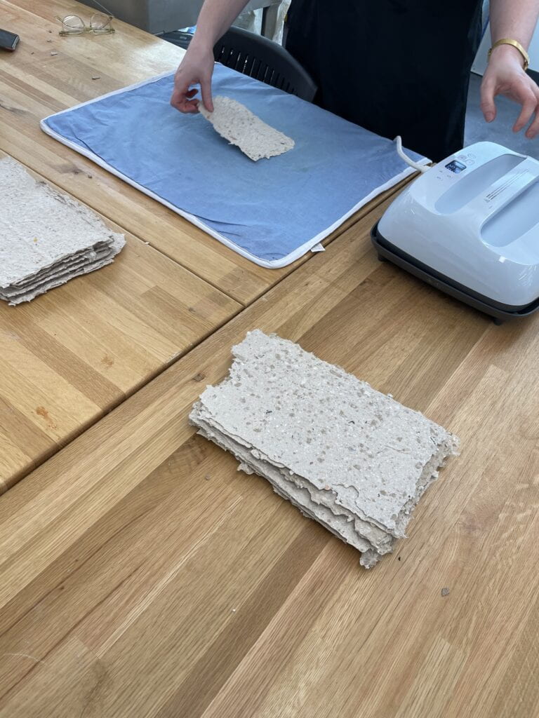 Image of wooden tabletop with a stack of handmade paper and hands ironing a sheet of paper 