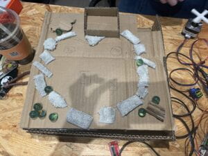 Cardboard box with a circle of fabric on top of it and green marbles in the corners.