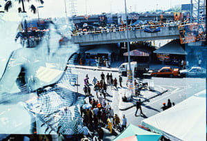 Streets lined with people and tents overlaid by the image of a woman laying in bed