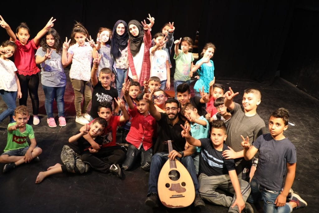 large group of children and man with guitar posing onstage giving peace sign