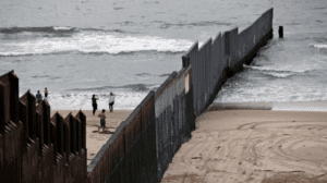 A photo of the wall that separates San Diego, California and Tijuana, Mexico, extending into the water of the two countries' shared coastline. There are people taking photos of the wall on the Tijuana side of the wall. 