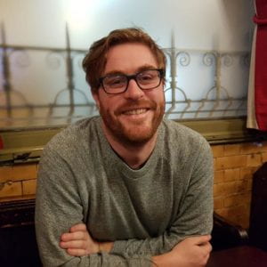 Red haired man with glasses smiling in front of a mural.