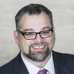 Man with rectangular black glasses smiles at the camera.