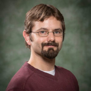 A man with red hair, a beard, and wire-framed glasses smiles at the camera