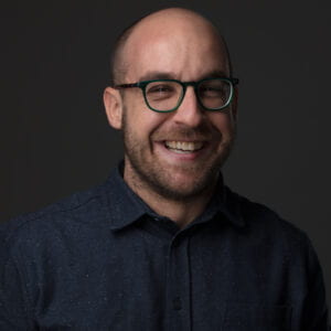 Man with glasses, and a beard smiles at camera against a black background.