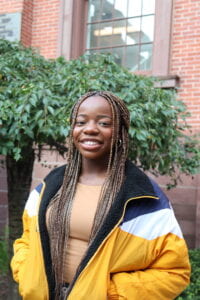 Dorothy Akpovwa smiling while wearing a yellow top and standing in front of a tree with a brick building behind it.