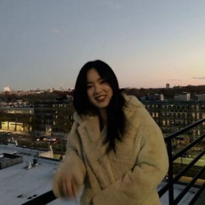 Jenny Zheng smiling while leaning on a balcony in front of a cityscape, during dusk.