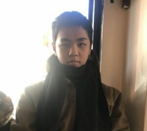 Eric Xu portrait, sitting in a corner, smiling in front of a window, wearing a brown coat and a black scarf.