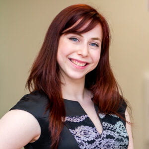 Avital smiling in front of tan background with red hair and blues eyes