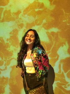 This is a Mihika, standing. She is smiling with her eyes closed and her hands behind her back. She is wearing a multicolored floral jacket and standing under yellow lighting.