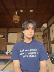 Photo of Matt Toups, a mixed white passing person with a black hair mullet, a pierced septum, and ears. They are wearing a blue shirt and sitting in a tatami room. 