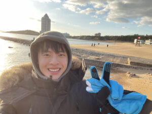 A picture of Sunny Qi in a black jacket, blue gloves, and a grey hoodie smiling.