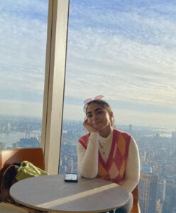 Krystine, a Latina woman, wearing a pink. orange, and white argyle sweater vest, sitting at a table.