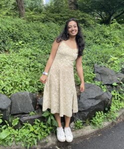 This is Ayushi. She is wearing a floral dress and has leaves and rocks in the background.