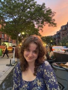 Bella sits down with an orange sunset behind them. They are wearing a purple blouse and have brown curly hair and bangs. They are smiling at their best friend who is taking the photo. 