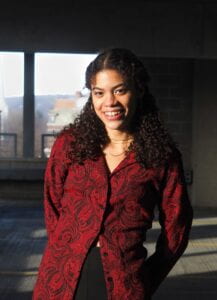 Kinana is standing in an industrial setting, with the image display her from the waist above. She is wearing a black pants with a red paisley patterned button down long sleeve. She has light brown colored skin with dark brown eyes, dark brown long curly and she is smiling showing teeth. 