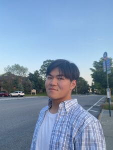 East Asian boy wearing a white shirt and a light blue flannel, while smiling at the camera and standing in front of a bus stop.