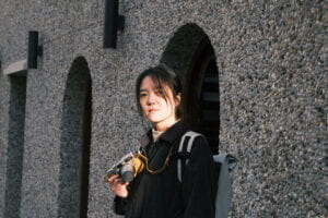 A picture of Muqing Wang holding film camera on her hand. Muqing is an asian female with black hair. She wears black coat and bag in this picture. 