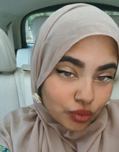 Brown female-presenting person with a brown hijab, facing the camera in selfie style with red lipstick and eyeliner