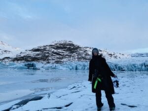 Me by Sólheimajökull glacier, wearing a grey beanie, a black coat with neon green safety belt on waist, black pants, white boots with black bottom on cramps, carrying a pair of white gloves on my left hand and a canon EOS camera on my neck, standing in the right 3/4 of the landscape.