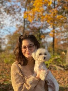 Female-presenting person with pink glasses facing the camera wearing woven turtleneck with a white, maltese dog to her right