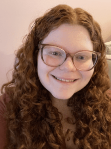 White woman with long, curly red hair, big pink glasses, and a maroon shirt. She is smiling and sitting in front of a light pink wall.