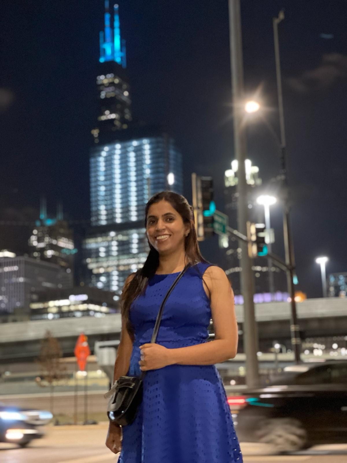 A South Asian woman with medium-length black hair, standing in a blue dress and smiling.