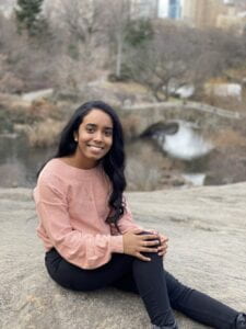 A brown female wearing a pink sweater and black jeans that is sitting on a rock in central park