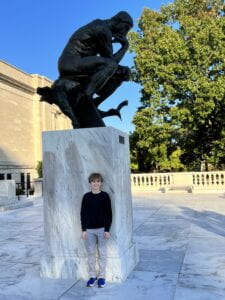 Boy standing in front of Rodan's The Thinker at the Cleveland Museum of Art outside on a sunny day.